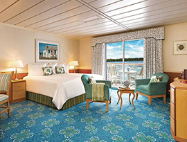 The Deluxe Balcony 1 stateroom on American Constitution small cruise ship, with floor-to-ceiling sliding glass doors that lead to a private balcony with a table and chairs overlooking the Inside Passage. There is a bed with a bedside table and a lamp on top of it on each side, and two chairs by the foot of the bed, all over a blue-patterned rug. The bed is facing a dresser and a TV on the wall. 