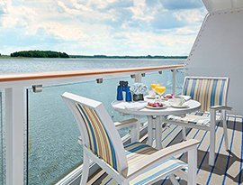 On the private balcony of American Constitution, an American Cruise Lines small cruise ship, two blue and yellow striped chairs surround a round, white table with a two plates of food and glasses of juice, binoculars, and a pot with blue flowers. Outside the glass railing is the Intracoastal Waterway.