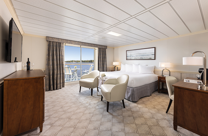 The spacious Sky Suite on American Constitution small cruise ship, with floor-to-ceiling sliding glass doors that lead to a private balcony with a table and chairs overlooking the Inside Passage. There is a bed with a bedside table and a lamp on top of it on each side, two chairs by the foot of the bed, and a painting of waves above it. The bed is facing a dresser with a TV on the wall above it.