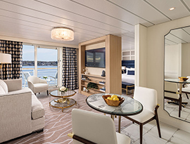 The Grand Suite on American Eagle, with floor-to-ceiling sliding glass doors that lead to a balcony with a table and chairs overlooking the Gulf Coast. There is a sofa facing a Tv stand with shelves and a TV on it, and a small, glass table between them. To the right, there is a round table with two white chairs and a large mirror on the wall. Behind the TV, there is a bed with dark blue pillows.
