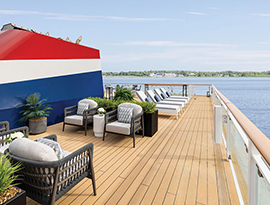 The sun deck on American Eagle, an American Cruise Lines Coastal Cat small cruise ship, with several large planters, and four plush outdoor arm chairs in front of four outdoor lounge chairs along beside the glass railings overlooking an intimate waterway.