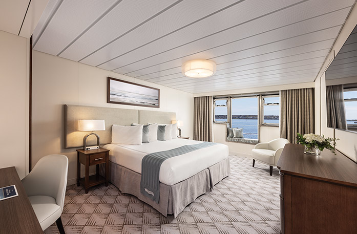 The Premium Waterview stateroom on American Eagle, with a large window in the center of two smaller windows, and a built-in-bench under them overlooking the Gulf Coast. There is a bed with white and grey pillows and a grey bed runner on the left side of the room, with a bedside table and a lamp on each side, and a painting of waves above it. The bed is facing a TV on the wall, above a dresser.