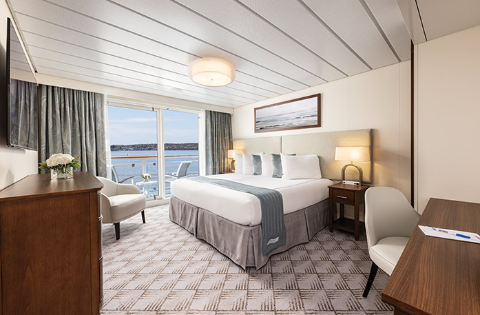 The Premium Balcony stateroom on American Eagle, with floor-to-ceiling sliding glass doors that lead to a balcony with a table and chairs overlooking the Gulf Coast. There is a bed with white and grey pillows and a grey bed runner on the right side of the room, with a bedside table and a lamp on each side, and a painting of waves above it. The bed is facing a TV on the wall, above a wooden dresser