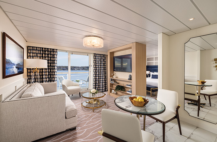 The Grand Suite on American Eagle, with floor-to-ceiling sliding glass doors that lead to a balcony with a table and chairs overlooking the Gulf Coast. There is a sofa facing a Tv stand with shelves and a TV on it, and a small, glass table between them. To the right, there is a round table with two white chairs and a large mirror on the wall. Behind the TV, there is a bed with dark blue pillows.