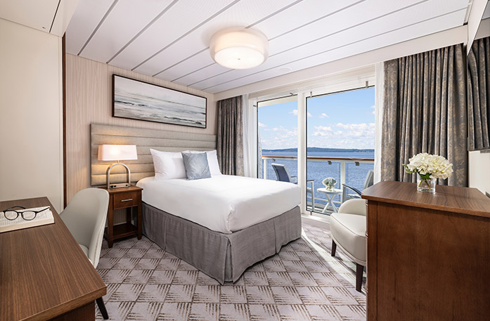 The Single Balcony stateroom on American Eagle, with floor-to-ceiling sliding glass doors that lead to a balcony with a table and chairs overlooking the Gulf Coast. here is a bed on the left side of the room with a grey accent pillow, a bedside table with a lamp, and a painting of waves above it. There is a wooden dresser on the right side, and a desk and chair in the foreground. 