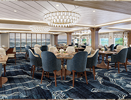 The restaurant on American Encore, where there are several round tables and navy blue chairs along the blue and white patterned carpet, and two round chandeliers hanging from the ceiling. There are large windows with clear views of the Columbia River on one wall, and floor-to-ceiling paintings on the back wall.