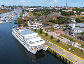 American Glory, an American Cruise Lines Coastal Cat small cruise ship docked in Brunswick, where there are many large buildings among trees and shrubs. In the distance, several small boats are also docked along the shoreline. 