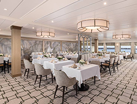 The Restaurant on American Glory, an American Cruise Lines Coastal Cat small cruise ship, where there are several plush chairs and various sizes of tables draped with white table cloths and perfectly set with folded napkins, glassware, and a vase with flowers in the center. 