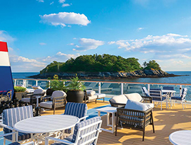 The sun deck on American Glory, an American Cruise Lines Coastal Cat small cruise ship, with several large planters and plush outdoor arm chairs, surrounded by glass railings around the edge of the deck. The ship is sailing by a small island covered in trees along the Maine coast.