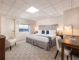 The Deluxe Balcony 2 stateroom on American Independence, an American Cruise Lines small ship with a push door leading to a private balcony, beside a large window overlooking the Chesapeake Bay. There is a bed on the right side of the room with an end table with a lamp on top on each side of the bed. Across from the bed, there is a cream-colored chair beside a wooden dresser, with a TV on the wall.
