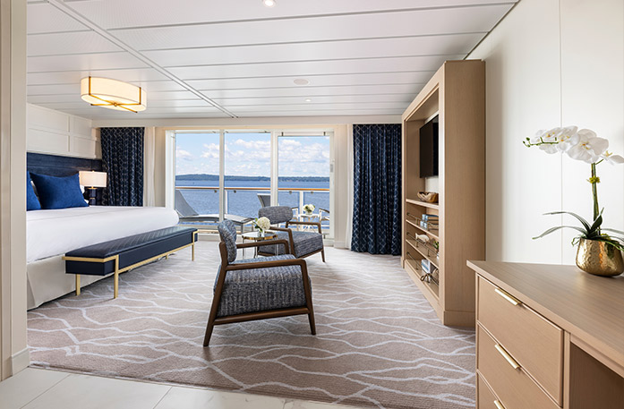 The Sky Suite on American Glory with floor-to-ceiling glass doors that lead to a balcony with a table and chairs overlooking the Gulf Coast. On the left side of the room, there is a bed with blue pillows and a blue bench with gold legs at the foot of the bed. There are two chairs with blue cushions in front  of a wooden TV stand with shelves and a TV on it. 