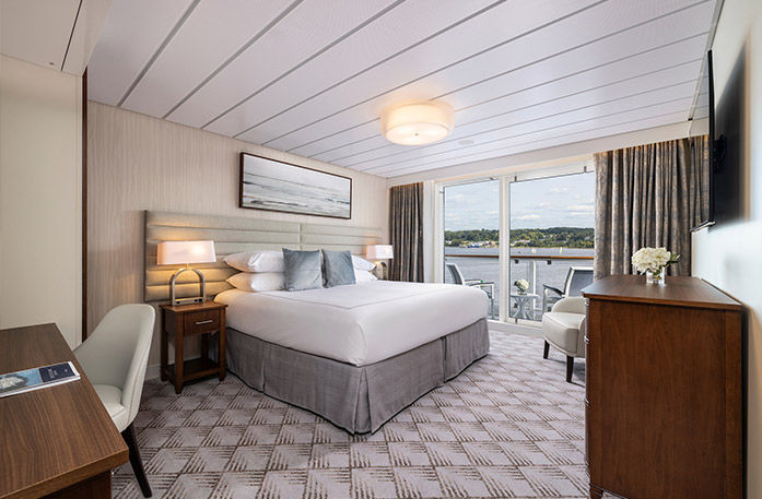 The Vista Balcony stateroom on American Glory with floor-to-ceiling glass doors that lead to a balcony with a table and chairs overlooking the Gulf Coast. There is a bed on the left side of the room with two grey pillows, bedside tables with a lamp on each side, and a painting of waves above it. There is a wooden dresser on the right side of the room with a TV on the wall above it. 