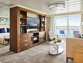 The Sky Suite on a American Harmony, with floor-to-ceiling glass doors that lead to a balcony with a table and chairs overlooking the Columbia River. There is a tan sofa with a black pillow beside a small table, and a large, wooden TV stand with six drawers and shelves filled with books and decor and a TV in the middle. Behind the tv stand is a bed with dark blue pillows with a painting above