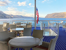 Several white chairs and tables cover the blue and white striped floor on the deck of American Harmony. There is a staircase leading to the deck below, and an American flag hung from a pole beside it. There is a view of the Columbia River with defined mountains on each side of the bright blue water.