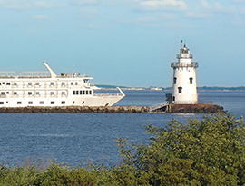 American Independence, an American Cruise Lines small ship cruising by the Saybrook Breakwater Lighthouse, a small white lighthouse in the middle of the water with a breakwater leading up to it, in Old Saybrook.