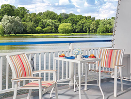 On the private balcony of American Independence, an American Cruise Lines small cruise ship, there are two chairs and a small table filled with two plates of food, a coffee mug, and a vase with blue flowers beside the white railing. The balcony provides a clear view of the Chesapeake Bay and the trees that line the shoreline. 