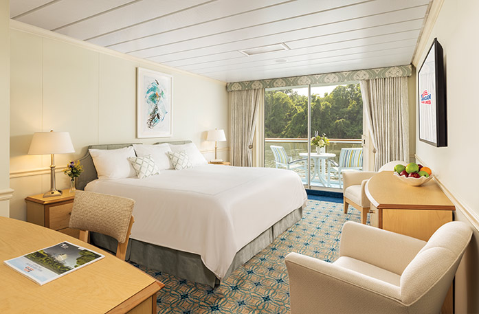 The Deluxe Balcony 1 stateroom on American Jazz with floor-to-ceiling glass doors that lead to a balcony with a table and chairs overlooking the Columbia River. There is a bed on the left side of the room with pillows and a silver bed frame. There are two bedside tables with lamps, two tan accent chairs with a wood dresser between them, a blue patterned rug, and a TV on the wall.