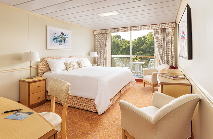The Premium Balcony stateroom on American Jazz with floor-to-ceiling glass doors that lead to a balcony with a table and chairs overlooking the Columbia River. There is a bed on the left of the room with pillows and a bed frame with an orange and white pattern. There are two end tables with lamps, two accent chairs with a wood dresser between them, an orange patterned rug, and a TV on the wall.
