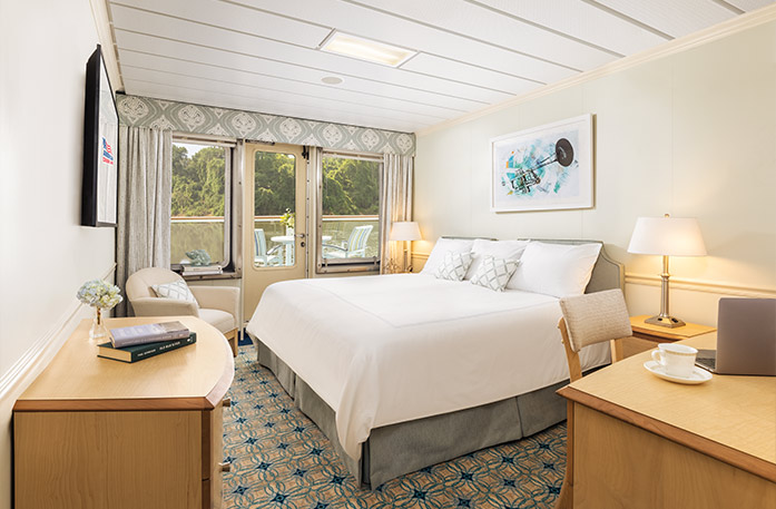The Standard Balcony stateroom on American Jazz with a push door that leads to a balcony with a table and chairs overlooking the Columbia River. There is a bed to the right with pillows and a grey bed frame. There are two end tables with lamps, an accent chair in the left corner, a wood dresser under a TV on the wall, and a desk and chair to the right of the bed.