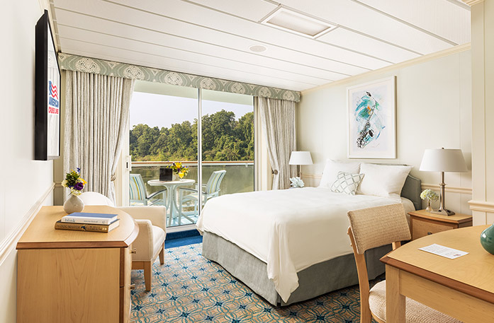 The Single Balcony stateroom on American Jazz with a push door that leads to a balcony with a table and chairs overlooking the Columbia River. There is a bed to the right with pillows and a grey bed frame. There are two end tables with lamps, an accent chair in the left corner, a wood dresser under a TV on the wall, and a desk and chair to the right of the bed.