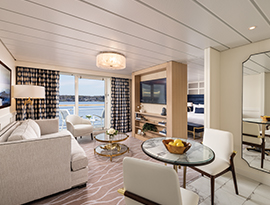 The Grand Suite on American Legend, with floor-to-ceiling sliding glass doors that lead to a balcony with a table and chairs overlooking the Gulf Coast. There is a sofa facing a TV stand with shelves and a TV on it, and a small, glass table between them. To the right, there is a table with two white chairs and a large mirror on the wall. Behind the TV, there is a bed with dark blue pillows.