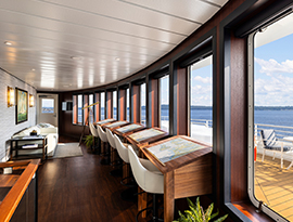 The Chart Room, a common area on American Legend Coastal Cat small cruise ship with five large maps on slanted wooden tables paired with white chairs against several large windows overlooking the sun deck with a table and chairs against the railing, and the Chesapeake Bay. 