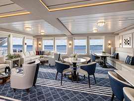The Sky Lounge, a common area on American Legend Coastal Cat small cruise ship with large, tan sofas along the walls with coffee tables in front of them, and a round table in the middle of the room with four blue chairs surrounding it. There are large windows along two of the walls, with views of the Chesapeake Bay.