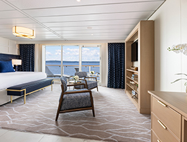 The Sky Suite on American Liberty with floor-to-ceiling sliding glass doors that lead to a balcony with a table and chairs overlooking the Gulf Coast. On the left side of the room, there is a bed with blue pillows and a blue bench with gold legs at the foot of the bed. There are two chairs with blue cushions in front  of a wooden TV stand with shelves and a TV on it. 