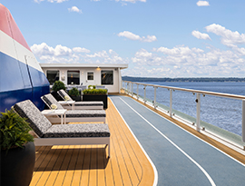 The sun deck on American Liberty, an American Cruise Lines Coastal Cat small cruise ship, with three outdoor lounge chairs, large potted plants, and the sky walk, a blue and white walking track surrounding the perimeter of the deck beside the glass railing with a view of the New England coast. 