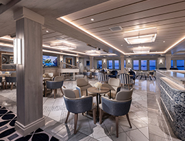 The Chesapeake Lounge, a common area on American Liberty Coastal Cat small cruise ship with several tables with four plush chairs surrounding them along the marble tiled floor. Windows line the back wall, with a view of the pink sunset.