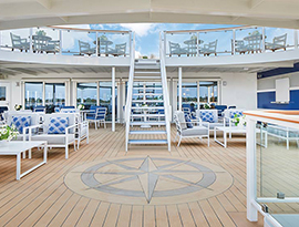 The sun deck of American Melody, featuring several grey outdoor lounge chairs with blue pillows and white square tables with a flower vase on top. In the center of the deck, there is a circle on the ground with a compass star engraved on it in front of a white and blue staircase leading up to the top deck with glass railings and more chairs and tables.