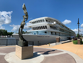 American Melody, an American Riverboat small cruise ship, docked in Red Wing. There is a light orange stone path leading up to a grey circle around The Balance of Power, a bronze statue with two eagles with their wings wide open in the air. In the distance, there are two American Cruise Lines tour buses.