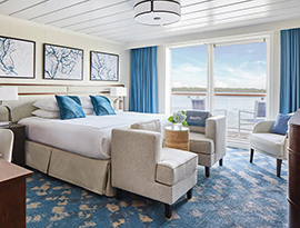 The Veranda Suite on American Melody, with large floor-to-ceiling glass doors, with blue curtains on each end, that lead to a balcony with a table and chairs overlooking the Mississippi River, a bed with white bedding and two blue pillows and two accent chairs at the foot of the bed, and three white paintings with blue lines above the bed. There is an end table with a lamp on each side of the bed.