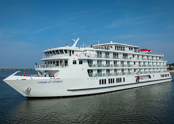 An aerial view of American Patriot, an American Cruise Lines Patriot Class small cruise ship sailing along the New England coast, with views of the shore in the distance.