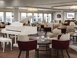 The Chesapeake Lounge, a common area on American Patriot with several couches, chairs and small round tables spread out among the burgundy, tan, and white carpet. There is a white grand piano in the center of the room, and floor-to-ceiling windows overlooking the New England coast.