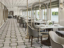 The Restaurant on American Patriot, where there are several individual dining tables surrounded by grey plush chairs in a line along a wall filled with large windows overlooking the New England coast.