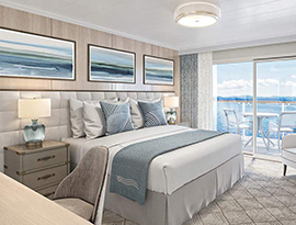 The Veranda Suite on American Ranger, with floor-to-ceiling sliding glass doors that lead to a private balcony overlooking the Hudson River. There is a bed against the wall on the left side of the room with a blue bed runner and accent pillows, an end table with a lamp on each side, and three paintings above the bed.