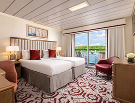 The Deluxe Balcony 1 stateroom on American Pride, with floor-to-ceiling sliding glass doors that lead to a private balcony with a table and chairs overlooking the Columbia River. There are two beds beside each other on the left side of the room with a red accent pillow on each, across from a wooden dresser and a TV on the wall above it across from the bed, and a red chair in the corner.
