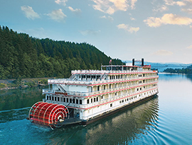 American Pride, an American Cruise Lines Paddlewheeler, sailing along the Columbia River. The back of the small cruise ship has a large, red paddlewheel, the shore on the left side of the river is covered in trees, and there is a tall bridge ahead in the distance.
