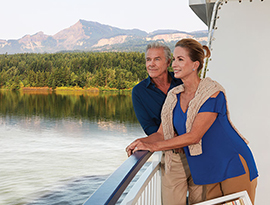 A couple leaning on the railing of their private balcony on American Pride, an American Cruise Lines Paddlewheeler, overlooking the Columbia River. In the distance, a silhouette of a mountain on the horizon. 