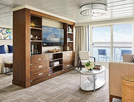The Grand Suite on American Serenade, with floor-to-ceiling glass doors that lead to a balcony with a table and chairs overlooking the Mississippi River. There is a tan sofa with a black pillow beside a small table, and a large, wooden TV stand with six drawers and shelves filled with books and decor and a TV in the middle. Behind the tv stand is a bed with dark blue pillows with a painting above.