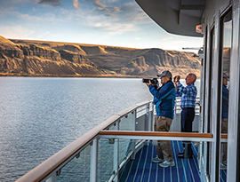 Two American Cruise Lines cruise guests taking photos of the scenic desert canyon mountain views from the deck of American Song on the Columbia River. One of the guests is taking a photo with a large camera, while the other is using his phone camera.