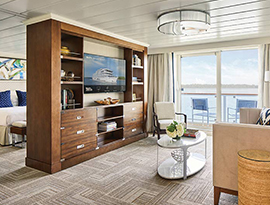 The Sky Suite on American Song, with floor-to-ceiling glass doors that lead to a balcony with a table and chairs overlooking the Columbia River. There is a tan sofa with a black pillow beside a small table, and a large, wooden TV stand with six drawers and shelves filled with books and decor and a TV in the middle. Behind the tv stand is a bed with dark blue pillows with a painting above