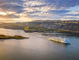 An aerial view of American Song, an American Riverboat small cruise ship sailing through the Columbia River Gorge, where there is a small island formation in the middle of the water. Alongside the water, there are several cliffs, and mountains in the distance.