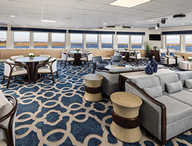 The Cascade Lounge, a common area on American Spirit, an American Cruise Lines small ship. There are several round tables with four chairs surrounding each, multiple grey couches, and windows along the walls with a view of Puget Sound.