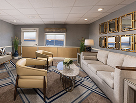 The Midship Lounge, a common area on American Spirit, an American Cruise Lines small cruise ship. There is a large, tan sofa on the right side of the room with gold-framed mirrors above it, two yellow arm chairs facing the sofa with a small, oval shaped table in the middle. There are two sofa benches against the wall on the other side of the room, and a large painting of sailboats on the wall 