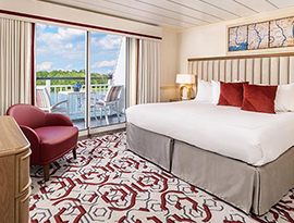 The Premium Balcony stateroom on American Splendor, with floor-to-ceiling sliding glass doors that lead to a private balcony with a table and chairs overlooking the Mississippi River. There is a bed on the right side of the room, and end tables with a lamp on each side of the bed. There is a dresser and a TV on the wall above it across from the bed, and a red chair in the corner.