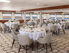 The Restaurant aboard American Star, an American Cruise Lines small cruise ship. There are several plush chairs surrounding round dinner tables draped with white table cloths and perfectly set with folded napkins, glassware, and a vase with flowers in the center