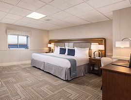 The spacious Veranda Suite on American Star, an American Cruise Lines small ship with a push door leading to a private balcony, beside a large window overlooking the Chesapeake Bay. There is a bed on the right side of the room with an end table with a lamp on top on each side of the bed. Across from the bed, there are two cream-colored chairs with a wooden dresser between them and a TV on the wall