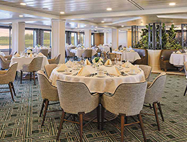 The restaurant on American Symphony, where there are several round tables covered with white tablecloths surrounded by grey plush chairs. There are perfectly folded napkins on the tables beside silverware and coffee mugs. There are windows along the left wall with views of the Mississippi River and the shoreline. 